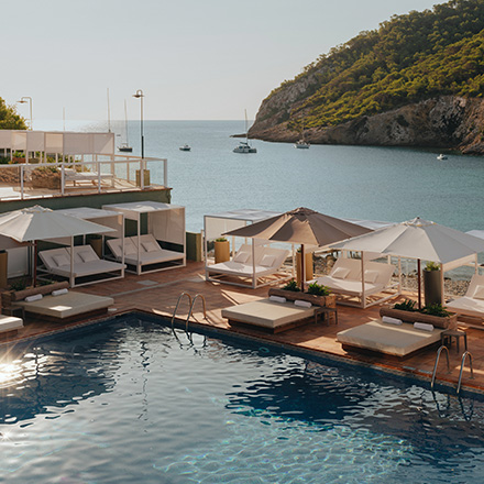 Piscina azul con grandes tumbonas y sombrillas de color crema en una zona de piscina con el mar azul y las montañas llenas de árboles al fondo.