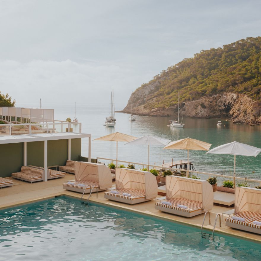 Piscine avec chaises longues sous un cabanon et parasols rayés, avec l'océan et les montagnes en arrière-plan