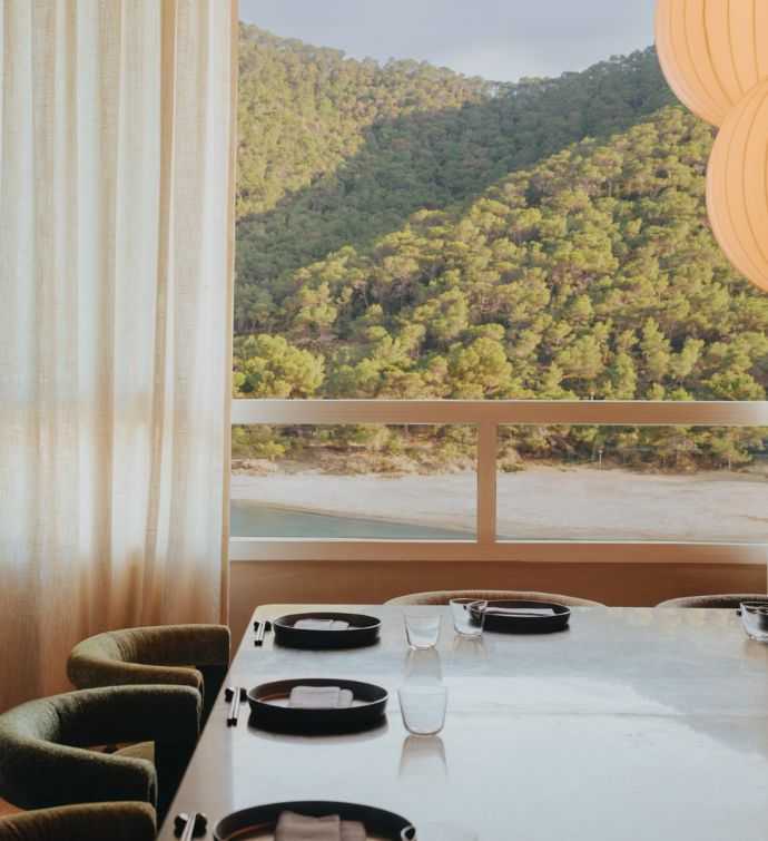 Corner of a large dining table set with plates with a curtained large window looking out to a beach and tree-covered mountains