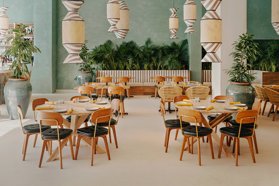 Dining room with light green walls, hanging lights with grey stripes, and round wooden tables and wooden chairs with green plants dispersed throughout.