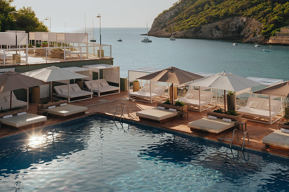 Large pool surrounded by beige chaise lounges and umbrellas with an ocean in the background with small boats floating and tree-filled mountains in the distance