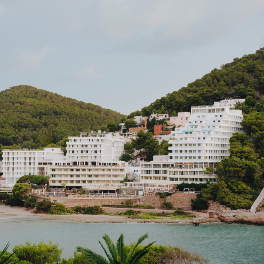 Gran edificio de hotel blanco en una playa costera de arena con montañas verdes al fondo