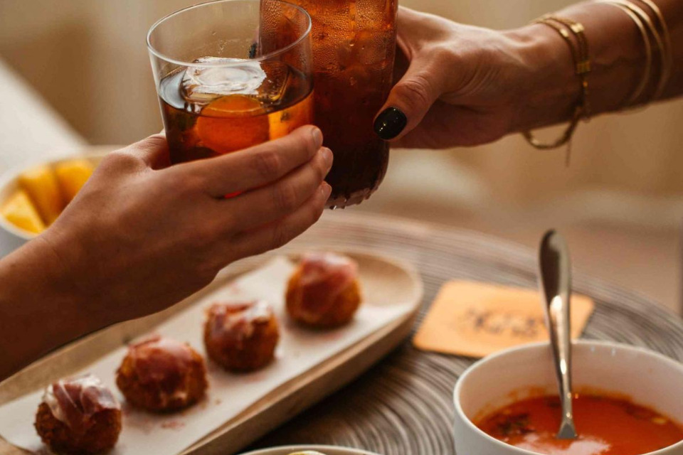 Two hands holding cocktails toast with croquetas and a soup on a table below them