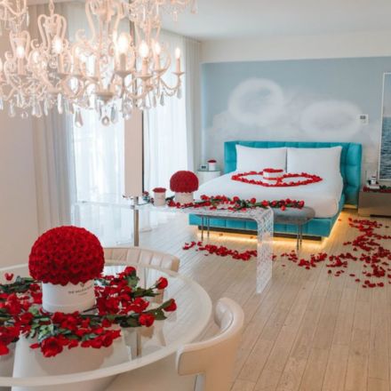 Large hotel suite with teal headboard bed with scattered rose petals all over it and the ground and a round glass table with a bouquet of roses and rose petals scattered about