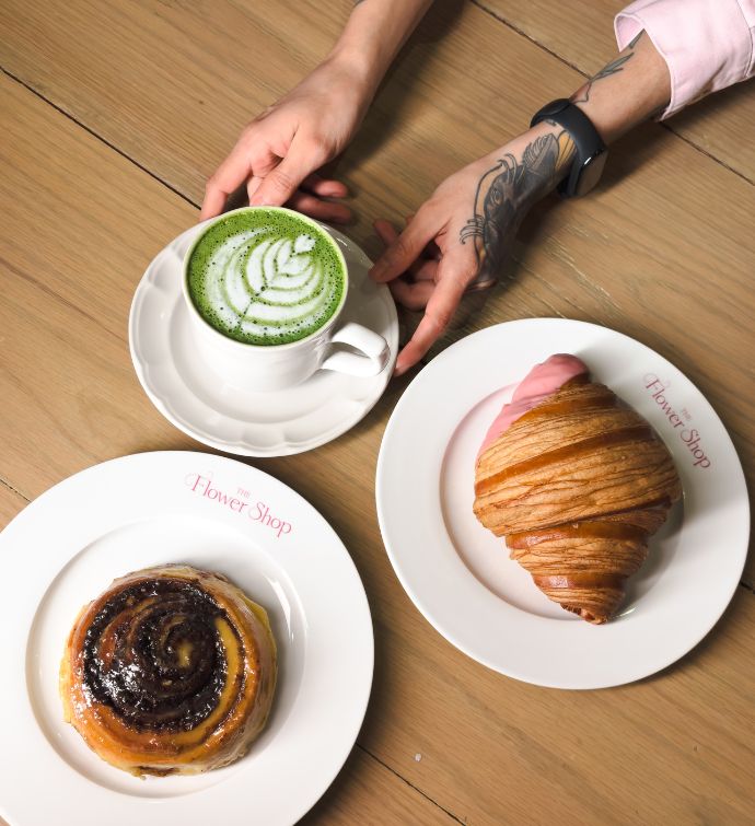 Croissant on a small white plate, bun on a small white plate, and a matcha latte with hands holding it