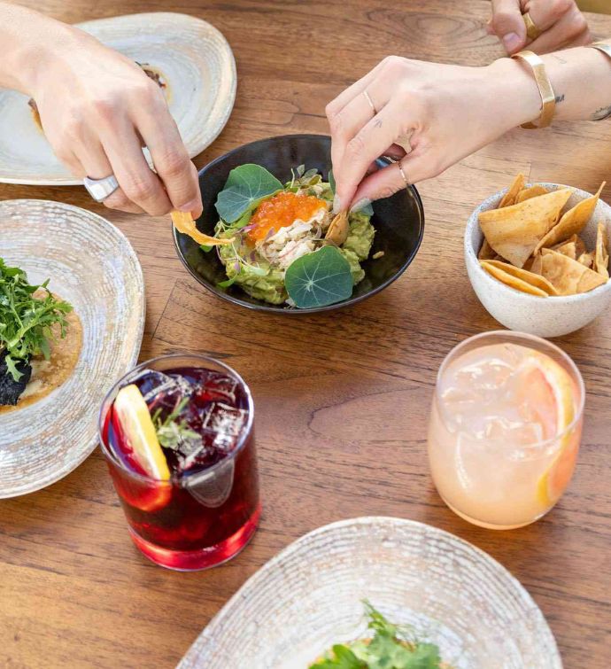 Small bowl of guacamole with two hands holding chips dipping into it with other dishes and two cocktails on the table