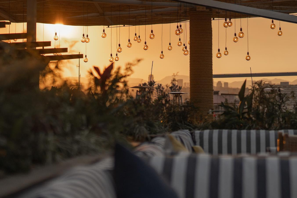 Terraza al aire libre con la puesta de sol al fondo, asientos de cojines a rayas en primer plano y techo del establecimiento.