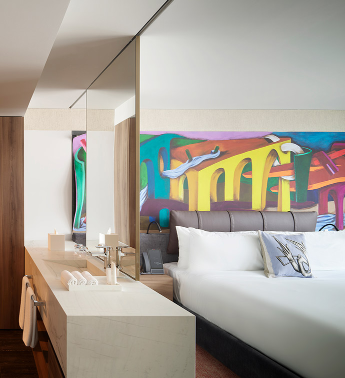 Hotel bed with a colorful wall mural, and bathroom with sink.