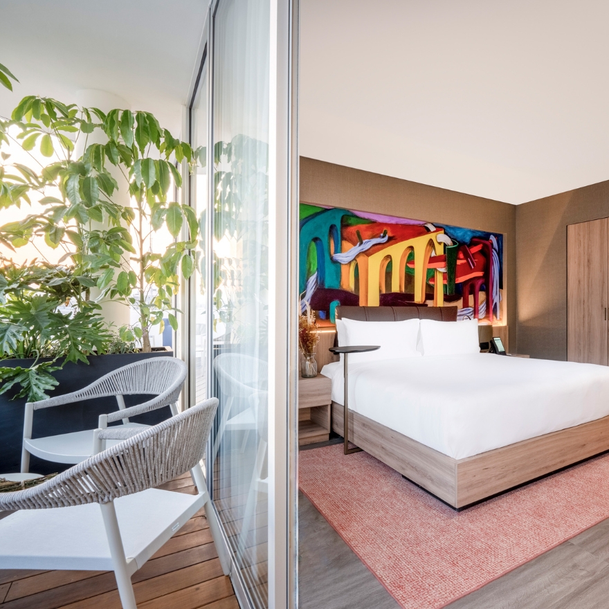 Hotel room with large white bed and colorful art piece on wall above and outdoor terrace with two white chairs and green plant