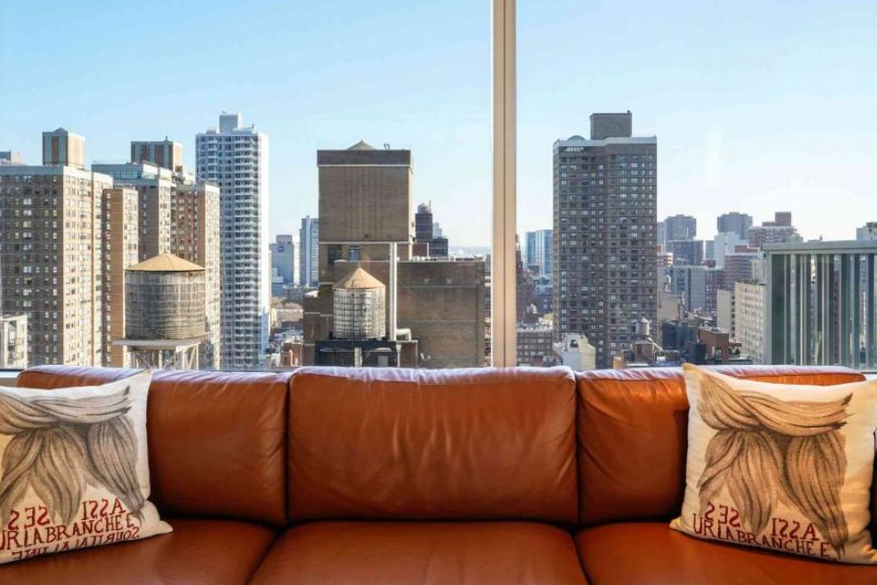 A brown leather couch with two pillows and views of New York out the window behind it