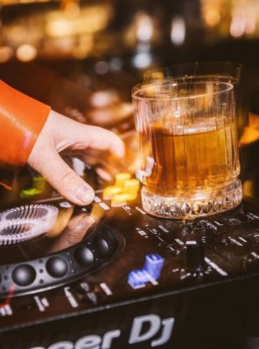 Glass of bourbon sits on top of a dj booth with a hand reaching towards it