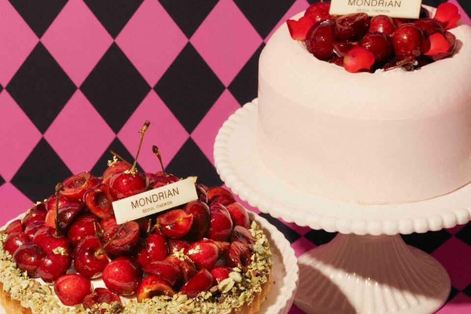 A pie with cherries on top and a white frosted cake with cherries on top sit on a surface with a black and pink checked background