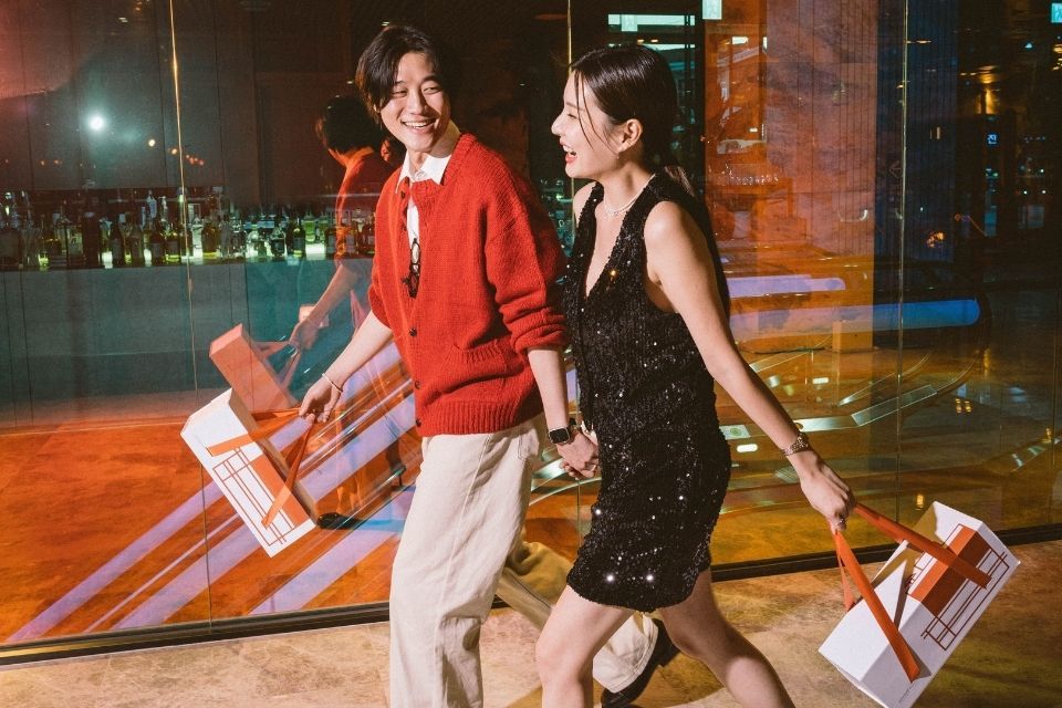 Man in red sweater and woman in black sparlky dress walk hand in hand with boxes in hand