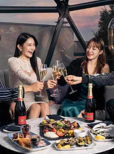 Two women sitting at a table with lots of food and toasting with champagne glasses