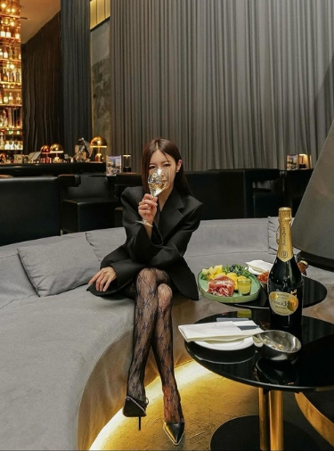 Woman in black dress sits on a grey sofa sipping a glass of champagne