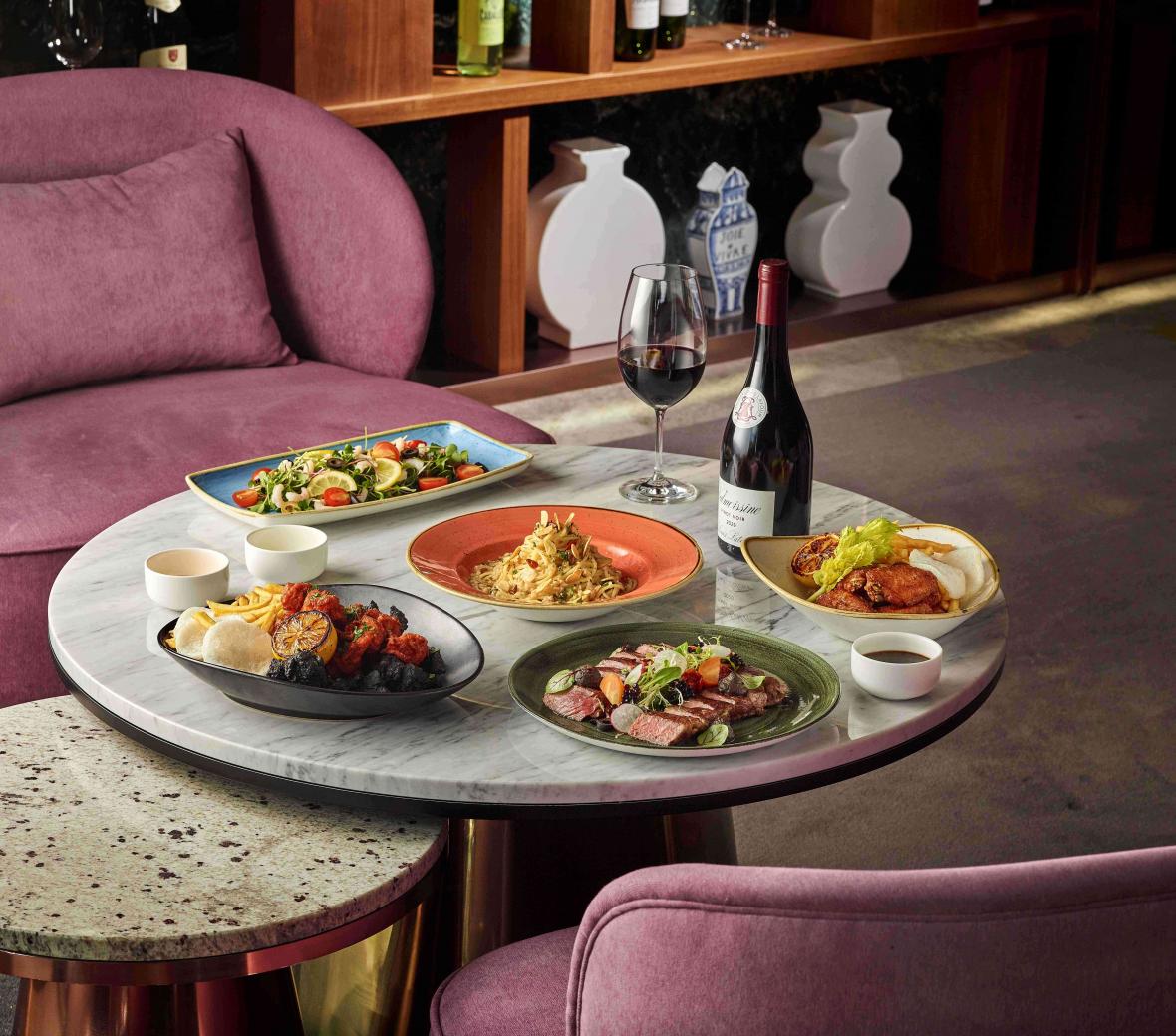 Multiple dishes and a bottle of wine sit on a round grey table with purple velvet lounge chairs