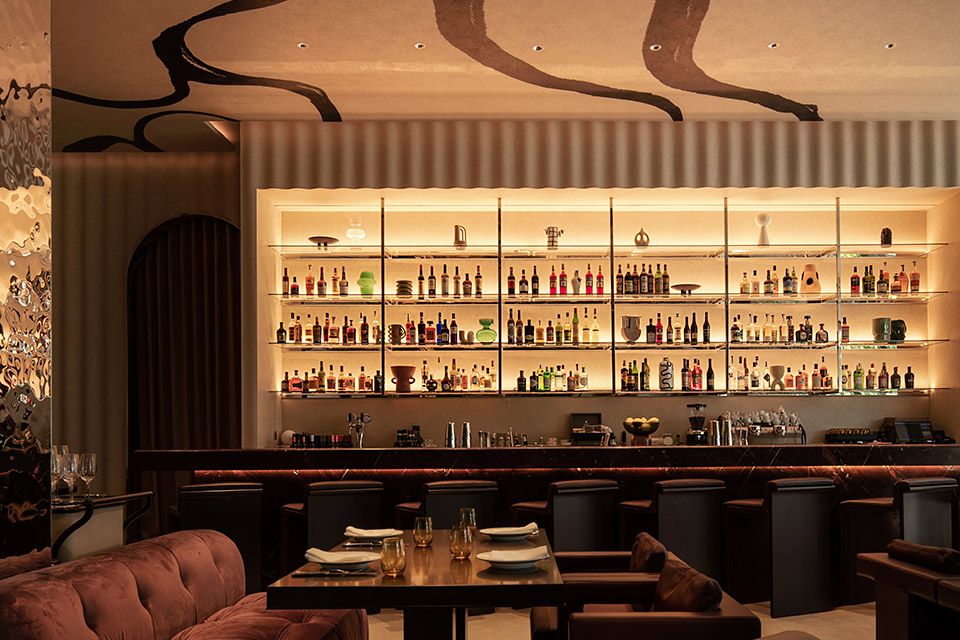 Dimly lit bar with shelves of liquor bottles, bar stools, and cushiony sofas, lounge chairs and a table in the corner.