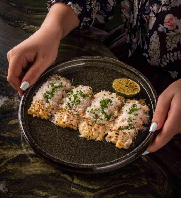 Corn with cheese and herbs on a dark round plate with hands holding it