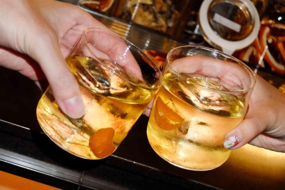 Close up image of two cocktails with orange slice floating in them held by two hands