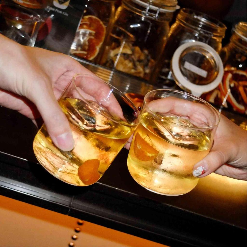 Two cocktails held by hands toasting in front of a bar