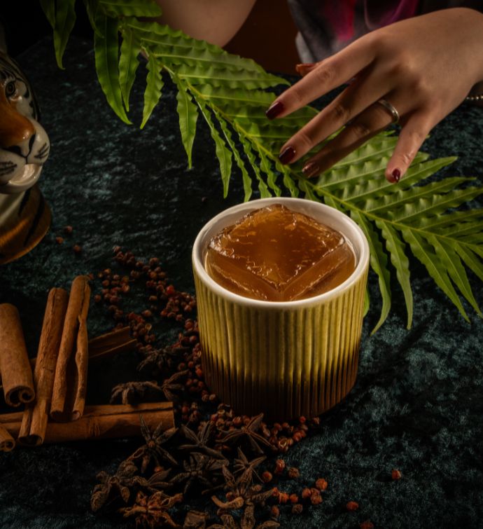 Small brown cocktail in a textured yellow cup on a table with palm leafs in the background and cinnamon sticks to the side