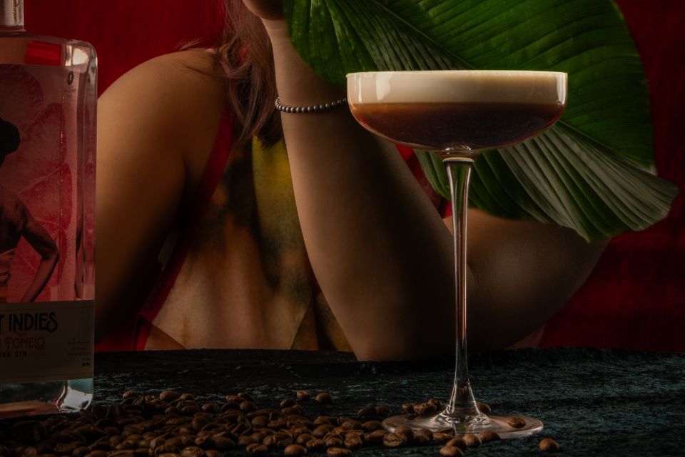 An espresso martini sitting on a dark bar top with big palm leaf and a woman's arm in the background