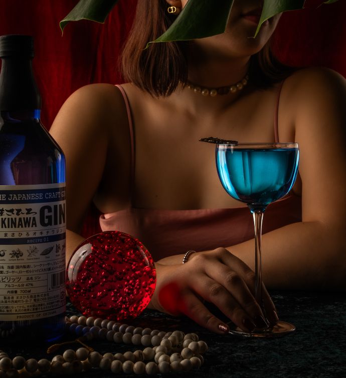 Bright blue cocktail with a woman holding it in the background and a bottle of gin to the side and a string of pearls on the table