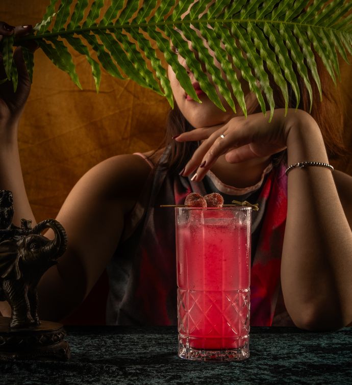 Bright hot pink cocktail in a tall glass with a womans arms in the background and a palm leaf covering her face