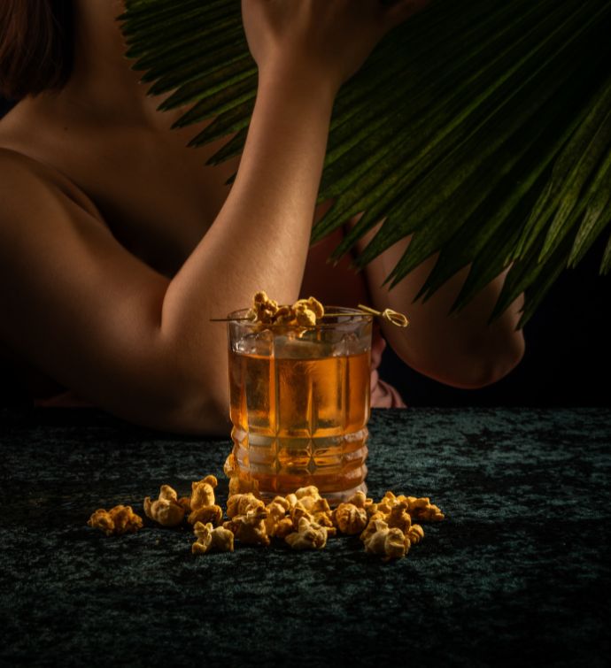 Light brown cocktail with garnishes on the table below and a woman's arm in the background with a palm leaf