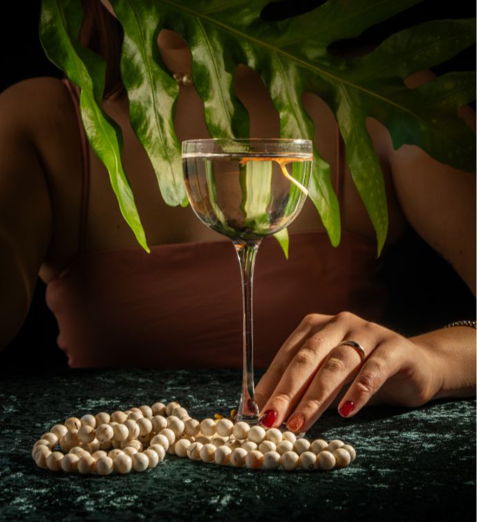 Clear cocktail in a small coupe glass with string of pearls on table and a woman's hand touching the stem of the glass