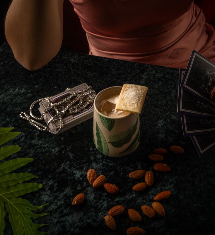 Light creamy cocktail with cracker garnish on a table with nuts scattered around a small silver purse on the side