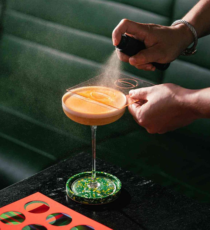 Cocktail in a coupe glass sits on a dark table while a bartender's hands sprays something over the cocktail.