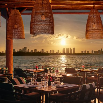 Beachfront deck with wooden tables, chairs, wicker pendant lights, and the sun setting in the distance over the ocean and city skyline