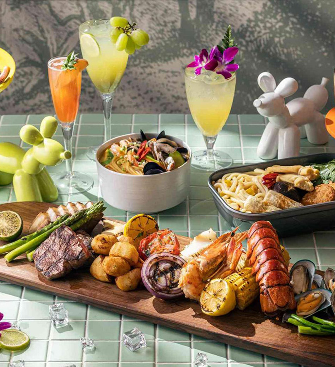 Spread of food and cocktails, including grilled lobster tail, steak, asparagus, corn and onions arranged on a board, alongside a seafood pasta