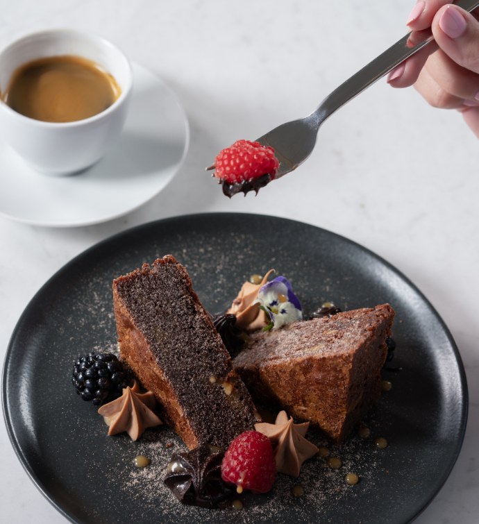 Tenedor levantando una frambuesa de un plato con un brownie rico en proteínas, acompañado de una taza de café