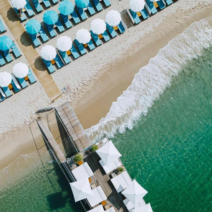 Vista aérea de una playa de arena con aguas turquesas y tumbonas con sombrillas blancas y azules