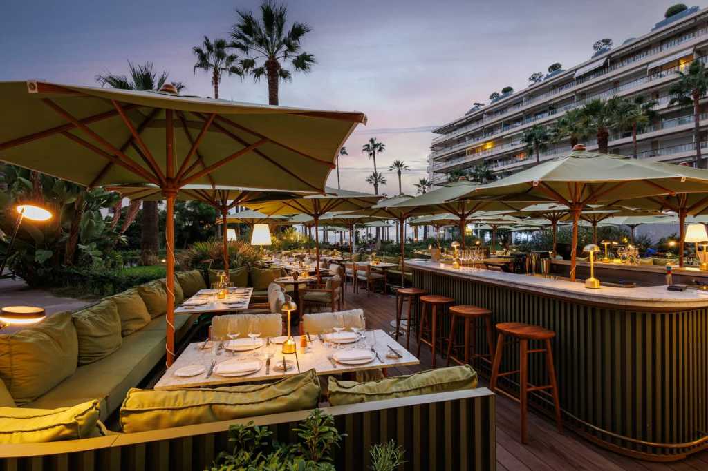 An outdoor dining patio in Cannes at dusk, with yellow umbrellas and palm trees in the distance