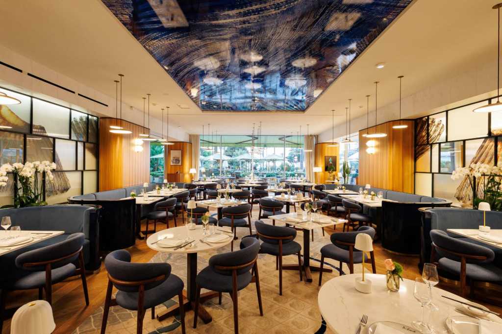 Large restaurant dining room with small cream-colored tablecloth tables with dark blue and wooden dining chairs, pendant lights hanging overhead, with a wooden floor with a large rug.