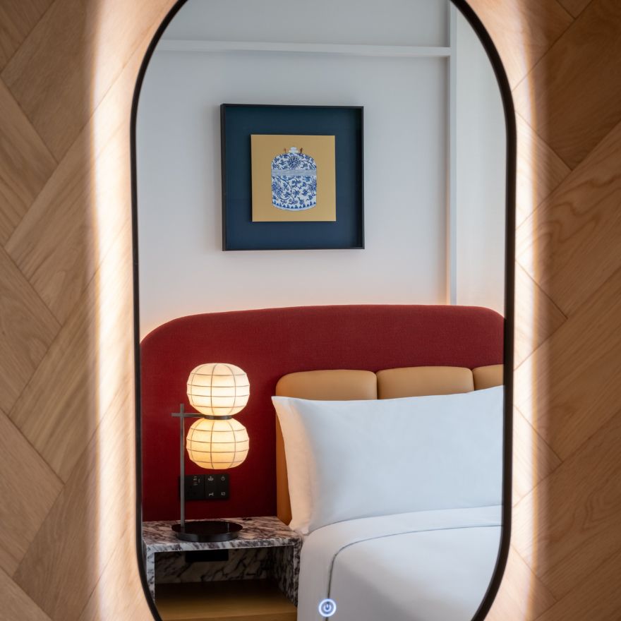 Large oval mirror with reflection of a bed with red headboard, small modern lamp on a tableside bed, and a framed piece of art on the wall above