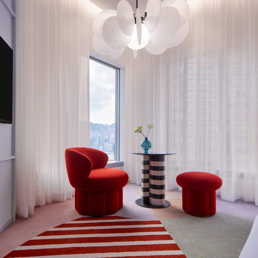 Room with long white curtains covering windows, a white decorative chandelier, small red lounge chair and stool, striped small table, and a striped red and white rug