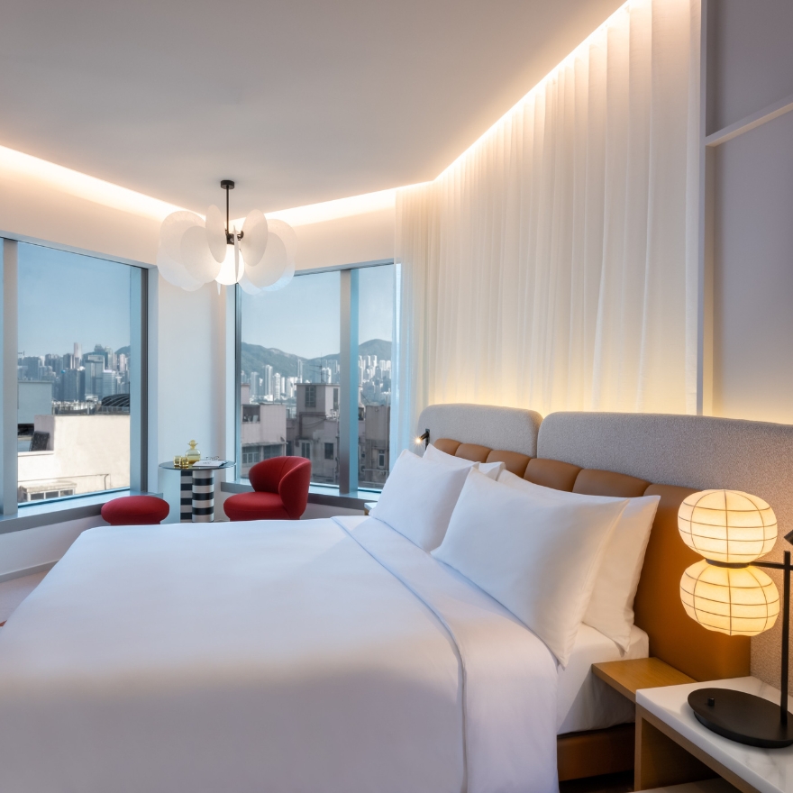 Large white bed with grey headboard and small lamp on a side table with a red lounge chair in the distance and large windows looking out to a city