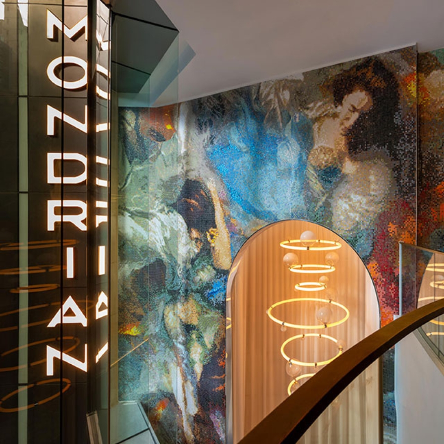 Hotel lobby view from a higher floor, with an illuminated Mondrian vertical logo on the left, a modern lit chandelier hanging from the ceiling, and a colorful mosaic mural on the wall.