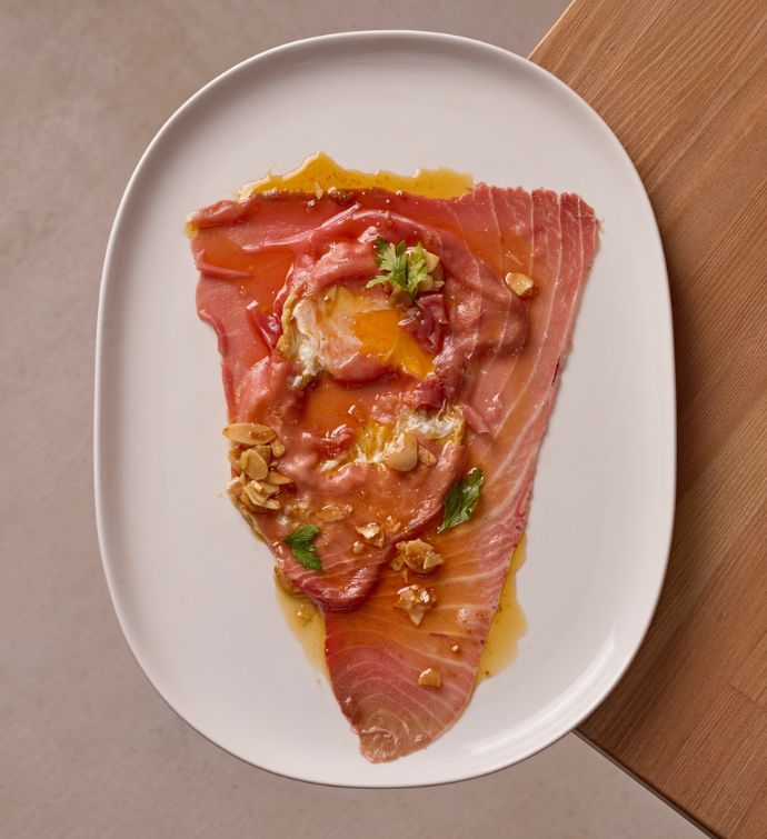 Fish carpaccio with garnishes on a white oval plate sitting on a light wooden table