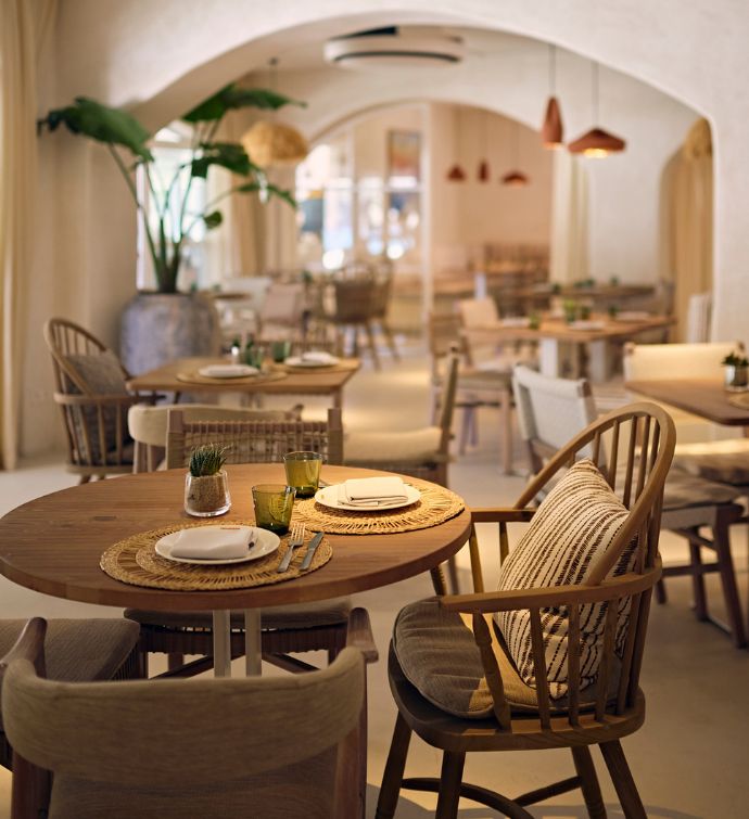 Bright dining room with circular light wooden table and mismatched chairs with plants and other tables in the background