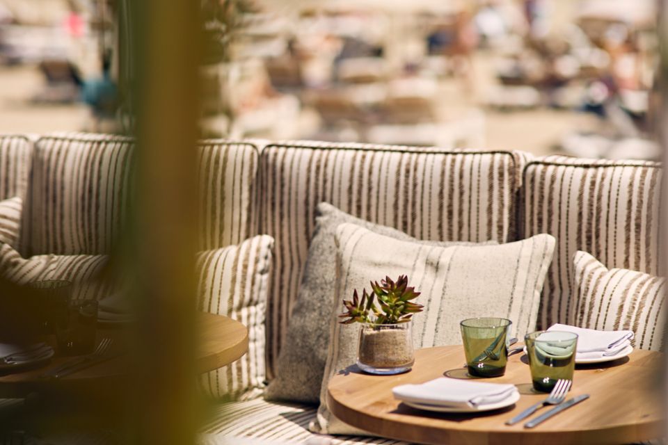 Close up image of a small wooden round table with plant, cups, and plates on top and a cushioned booth with extra striped pillows behind it and a beach blurred in the background