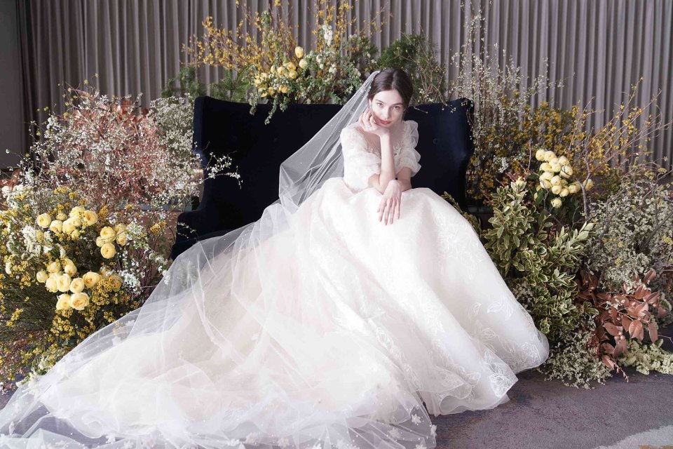 Woman in white wedding gown sits down with head resting on hands surrounded by big bouquets of flowers