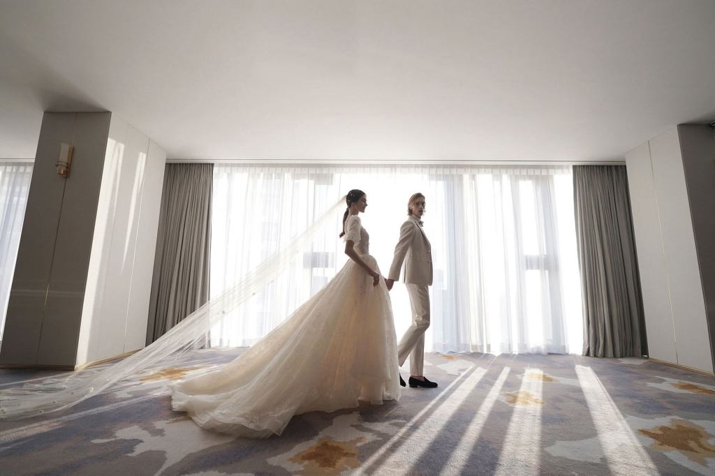 Bride in white gown and veil holds hands with groom in light cream suit while they walk past a window