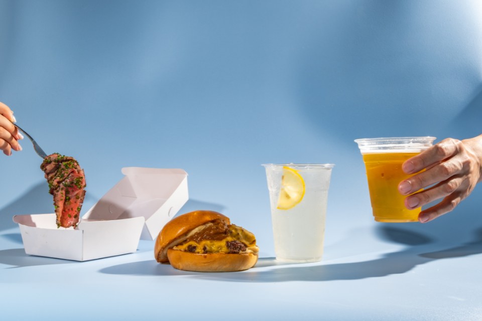 A burger, glass of lemonade, a hand holding a small beer, and a fork lifting up a piece of meat on a blue background