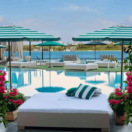 Cama junto a la piscina con almohadas a rayas verdes y blancas, rodeada de flores rosas, con la bahía de Biscayne al fondo.