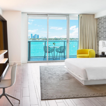 a large room with a rug, made bed, desk, yellow chair and balcony that overlooks the bay